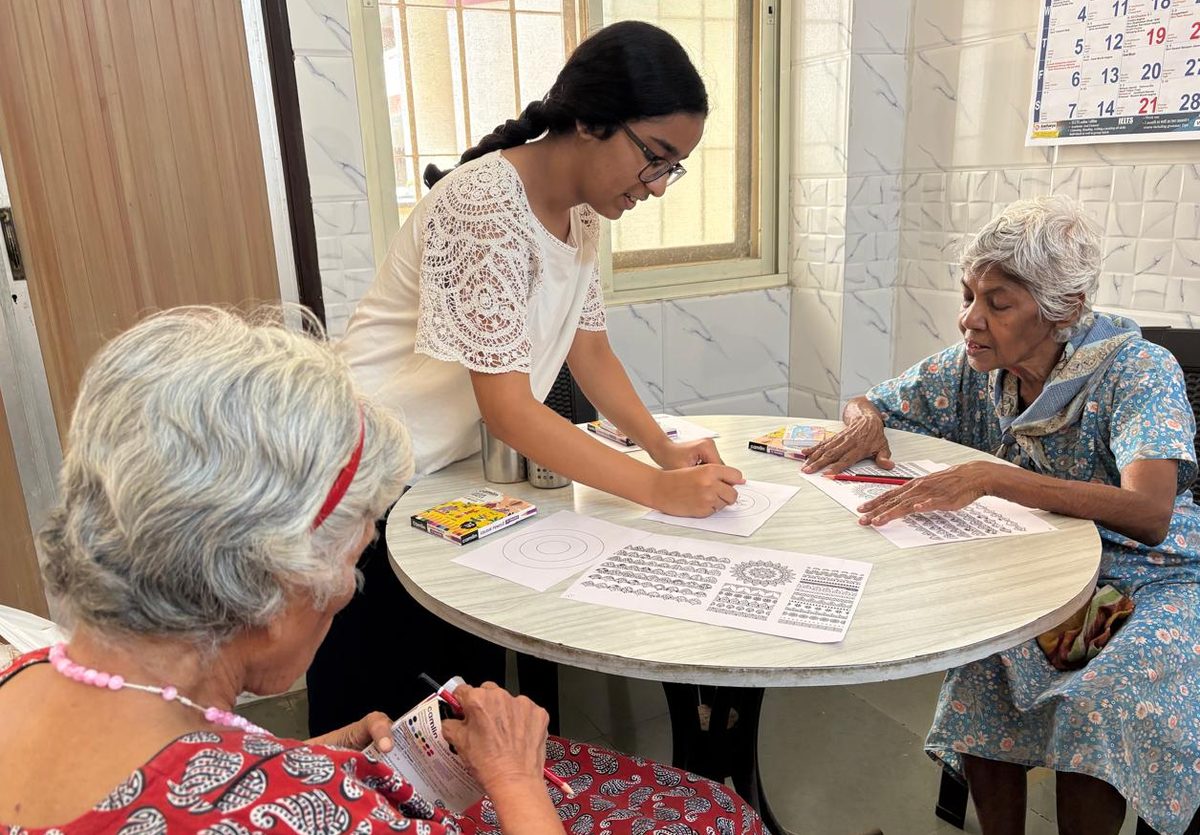A volunteer facilitator leaning in to help two older participants seated at a round table covered with worksheets and colouring materials.